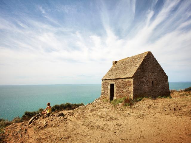 Une randonneuse assise à côté de la cabane Vauban de Carolles sur la falaise admire la baie du Mont Saint-Michel