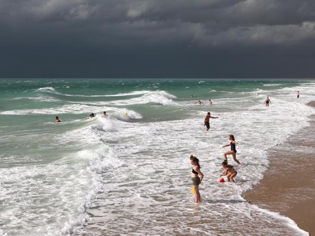 Des gens se baignent dans les eaux vertes et bleues de la mer à la plage de Saint-Martin-de-Bréhal sous un ciel gris et orageux